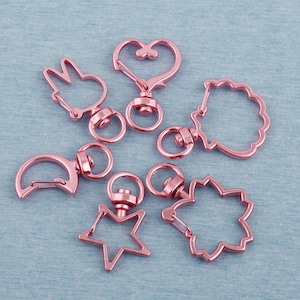 May include: A set of seven rose gold metal keychains with different shapes, including a heart, a star, a crescent moon, a bunny, and a flower. The keychains are all attached to swivel clasps.