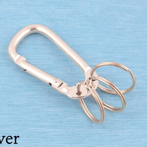 May include: A silver-toned metal carabiner keychain featuring a spring-loaded gate mechanism and three attached circular split rings for holding keys. The hardware has a smooth, polished finish and is displayed against a light blue background.