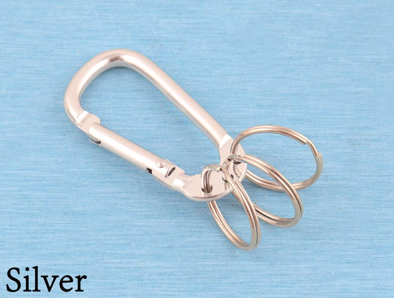 May include: A silver-toned metal carabiner keychain featuring a spring-loaded gate mechanism and three attached circular split rings for holding keys. The hardware has a smooth, polished finish and is displayed against a light blue background.