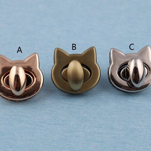 Op de afbeelding: Drie metalen kattenvormige sluitingen in verschillende afwerkingen: goud, brons en zilver. Elke sluiting heeft een rond, verhoogd oog in het midden.