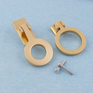 May include: Two gold-colored cabinet pulls with a circular design and a rectangular backplate. A small screw is included. The pulls are made of metal and are set against a light blue background. These are suitable for furniture restoration or new builds.