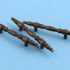May include: Three antique brass drawer pulls with a spindle-shaped design. Each handle features a central bar with decorative ribbed detailing and two mounting posts on the underside, finished in a dark, weathered metallic tone throughout.