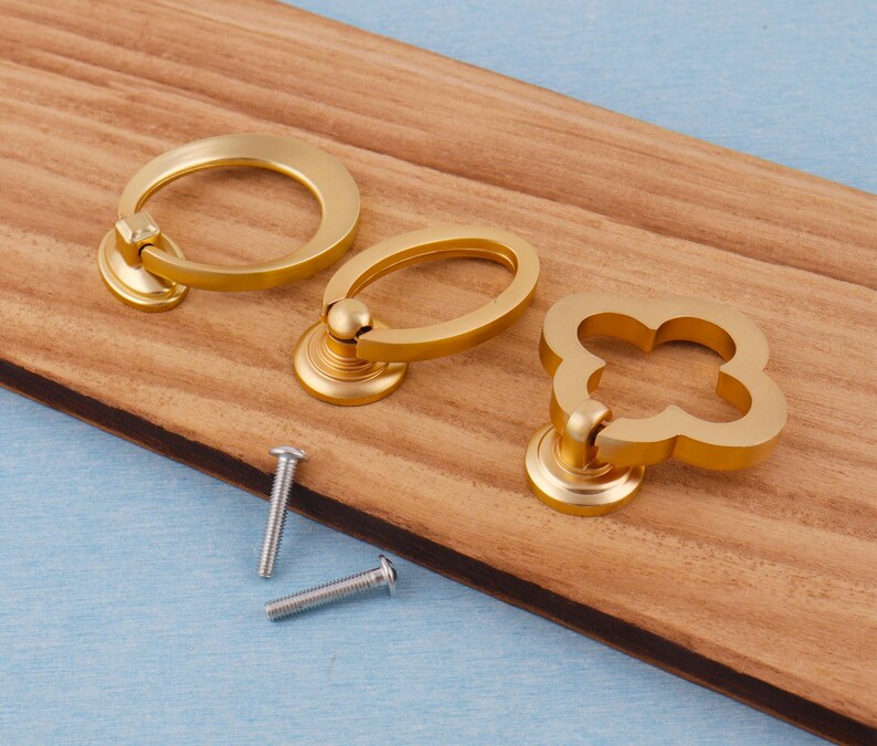 Może przedstawiać: Trzy złote uchwyty do szafek o r&oacute;żnych kształtach: pierścień, owal i czterolistna koniczyna. Widoczne są r&oacute;wnież dwie srebrne śruby. Uchwyty znajdują się na drewnianej powierzchni z niebieskim tłem.