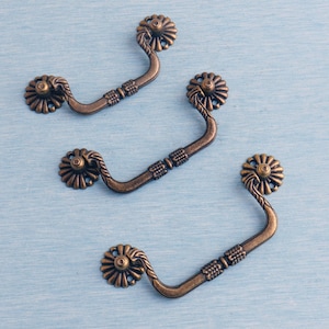 Könnte beinhalten: Vier antike Schubladengriffe aus Bronze mit floralem Design. Jeder Griff hat eine dekorative blütenförmige Rückplatte und einen gebogenen Griff mit seilartiger Textur. Die Griffe liegen auf einer hellblauen Holzoberfläche.