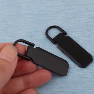 K&ouml;nnte beinhalten: Zwei schwarze Metall-Rei&szlig;verschlussanh&auml;nger mit rechteckigem Design und gebogenem B&uuml;gel. Einer wird von einer Hand gehalten, der andere liegt auf einer hellblauen Oberfl&auml;che. Die Anh&auml;nger haben ein mattes Finish.