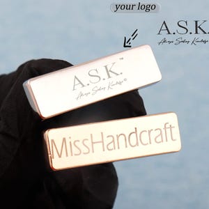 Può includere: Sono presentati due oggetti metallici rettangolari. Quello superiore è argentato con il testo "A.S.K. Always Seeking Knowledge" inciso. Quello inferiore è oro rosa con "MissHandcraft" inciso. Lo sfondo è azzurro.