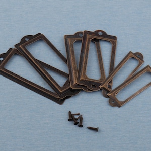 Puede incluir: Una colección de portatarjetas de color bronce antiguo con marcos rectangulares y tapas redondeadas. Varios tornillos pequeños están dispersos cerca. Los portatarjetas son probablemente para cajones o armarios.