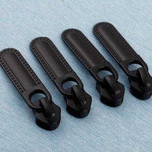 May include: Four black zipper pulls with a decorative beaded edge. The pulls are made of metal and have a rectangular shape.