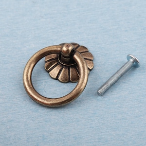 Op de afbeelding: Antieke messing ringgreep met een decoratieve bloemenachterplaat en een zilveren schroef. De meubelbeslag is een warme, verouderde bronzen kleur. De ringgreep is voor kasten.