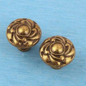 Könnte beinhalten: Zwei runde Schrankknöpfe mit einer metallischen Oberfläche in Antikmessing. Jeder Knopf weist ein erhabenes, wirbelndes, blumenartiges Muster auf, das von einem glatten, kreisförmigen Mittelpunkt ausgeht und eine texturierte Oberfläche erzeugt.