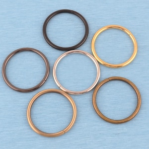 May include: A collection of six circular metal rings arranged in a cluster. The rings feature various metallic finishes, including polished gold, matte black, brushed bronze, copper, and silver tones, each with a visible seam along the circular band.