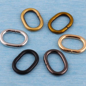 Puede incluir: Seis anillos metálicos ovalados en varios acabados: plateado, dorado, bronce y negro. Cada anillo tiene un diseño con bisagras, lo que permite abrirlo y cerrarlo. Los anillos están dispuestos sobre una superficie azul claro.