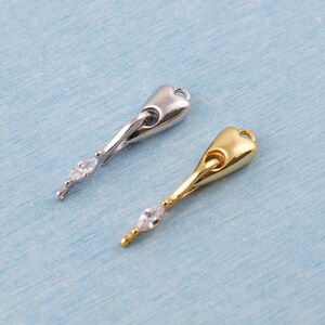 May include: Two pendant charms, one silver-toned and one gold-toned, each featuring a teardrop-shaped top and a slender, curved stem. A clear, marquise-cut gemstone adorns the bottom of each charm, adding a touch of sparkle. The charms are designed for jewelry making.