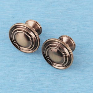 May include: Two round cabinet knobs with a brushed antique bronze finish. Each knob features a raised centre dome surrounded by concentric circular ridges, creating a tiered profile that tapers down to a narrow cylindrical base for mounting.