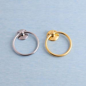 Pode incluir: Duas puxadores de anel de metal, uma prateada e outra dourada, são exibidas contra um fundo azul claro. Cada puxador apresenta um anel circular preso a uma base redonda. O puxador prateado está à esquerda e o dourado à direita.