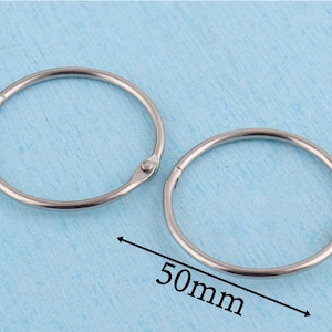 May include: Two circular silver-toned metal binder rings with a hinged opening mechanism. The rings are smooth and polished. A black arrow and the text 50mm are printed on the image, indicating the diameter of the metal rings is 50 millimeters.