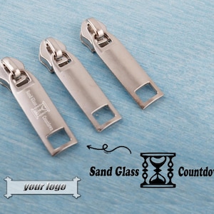 Op de afbeelding: Drie zilveren ritstrekkers met een rechthoekig ontwerp en een vierkante opening aan de onderkant. Eén trekker heeft de tekst "Sand Glass Countdown" erop gedrukt. De achtergrond is lichtblauw.