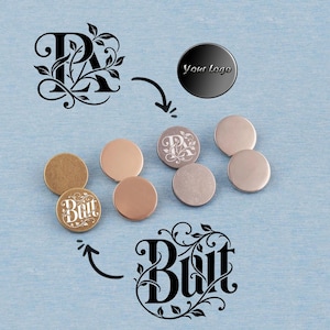 Könnte beinhalten: Eine Sammlung runder Metallknöpfe in Gold- und Silbertönen. Einige Knöpfe sind mit Designs graviert, darunter der Buchstabe "R" mit floralen Akzenten und das Wort "Butt". Ein schwarzer Knopf zeigt den Text "Your Logo".