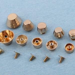 May include: A collection of gold-toned metal furniture feet and screws. The feet vary in shape, including cone and rounded designs, with textured and smooth surfaces. The screws are small with flat heads. The items are arranged on a light blue surface.