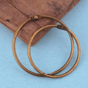 May include: Two circular metal binder rings with an antique bronze finish. Each ring features a hinged mechanism with a small rivet, allowing the hoop to open and close securely. The rings are constructed from a thin, rounded metal wire material.