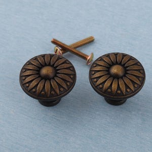 Puede incluir: Dos tiradores de armario de bronce antiguo con diseño de girasol. Los tiradores son redondos con un patrón detallado de pétalos y un acento circular central. Se incluyen dos tornillos dorados.