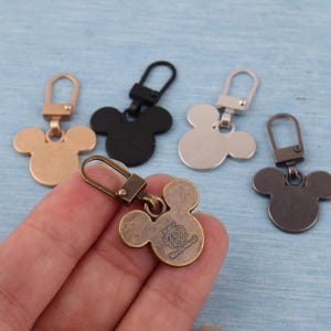 Może przedstawiać: Pięć metalowych zawieszek do zamków błyskawicznych w kształcie Myszki Miki w odcieniach złota, czerni, srebra i brązu. Jedna zawieszka jest trzymana w dłoni, pokazując tył z logo. Zawieszki mają zapięcie na karabińczyk ułatwiające mocowanie.