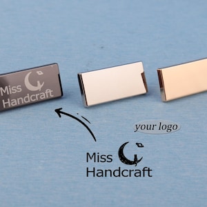 May include: Three rectangular metal name badges. One is black with the text "Miss Handcraft" and a crescent moon logo. The other two are silver and gold. The image also includes the text "your logo" and an arrow pointing to the "Miss Handcraft" logo.