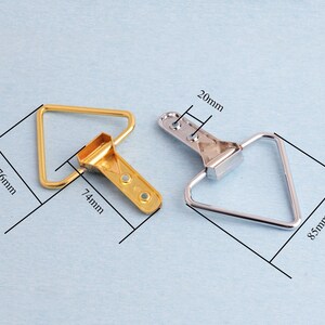 May include: Two metal picture frame hangers. One is gold-coloured, measuring 7.4cm x 7.6cm, and the other is silver-coloured, measuring 8.5cm. Both have a triangular loop and a bracket with holes for mounting. The bracket is 2cm wide.