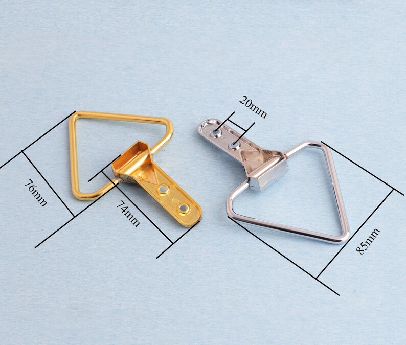 May include: Two metal picture frame hangers. One is gold-coloured, measuring 7.4cm x 7.6cm, and the other is silver-coloured, measuring 8.5cm. Both have a triangular loop and a bracket with holes for mounting. The bracket is 2cm wide.
