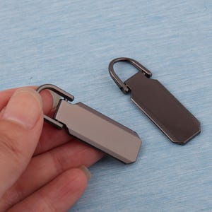 K&ouml;nnte beinhalten: Zwei Rei&szlig;verschluss-Zipper in Gunmetal-Farbe mit rechteckigem Design und gebogenem Griff. Einer wird von einer Hand gehalten, der andere liegt flach. Die Zipper haben ein gl&auml;nzendes, metallisches Finish auf hellblauem Hintergrund.