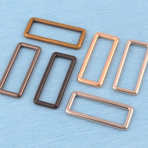 Pode incluir: Um conjunto de seis argolas retangulares de metal com vários acabamentos metálicos, incluindo latão antigo, castanho escuro mate, ouro rosa polido e prata. Cada argola apresenta um design de estrutura arredondada e lisa com centro aberto.