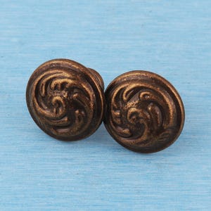 May include: Two round metal buttons featuring a raised, swirling embossed pattern in a dark bronze finish. The buttons have a circular shape with a textured, antique-style surface detail that creates a sense of depth across the face of each piece.