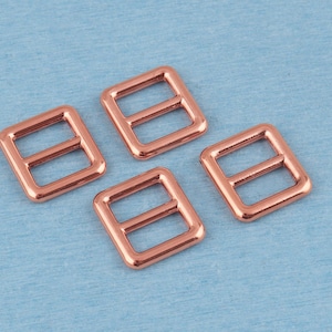 Puede incluir: Cuatro hebillas deslizantes rectangulares de metal con un acabado pulido en oro rosa. Cada hebilla presenta un marco liso y redondeado con una barra horizontal central, diseñada para ajustar correas en bolsos, prendas o accesorios.