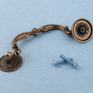 Può includere: Maniglia per cassetto in bronzo in stile antico con dettagli decorativi e due viti blu. La maniglia presenta un'impugnatura curva e terminali circolari con motivi intricati, ideale per il restauro di mobili o progetti fai-da-te.