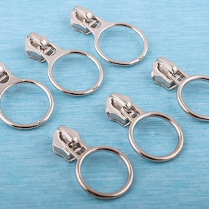 May include: A set of six silver-toned metal zipper pulls, each featuring a circular ring attachment. The pulls have a polished finish and are designed for attachment to zipper sliders, providing a round handle for easier gripping and operation.