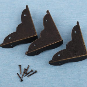 Może przedstawiać: Trzy antyczne, brązowe kątowniki z dekoracyjnymi wzorami kwiatowymi i małymi otworami na śruby. Obecnych jest również kilka małych, brązowych gwoździ, co sugeruje zestaw do obróbki drewna lub naprawy mebli.