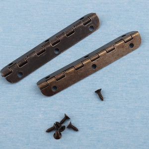 Op de afbeelding: Twee antiek bronzen scharnieren met meerdere segmenten en schroefgaten worden getoond. Verschillende kleine, donkere schroeven liggen in de buurt verspreid. De scharnieren zijn ontworpen om twee oppervlakken te verbinden.
