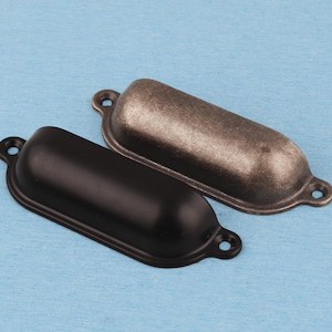 May include: Two metal cup drawer pulls, one with a matte black finish and the other with a distressed antique bronze finish. Each handle features a rounded, elongated shape with two small circular mounting tabs on either side for screw attachment.