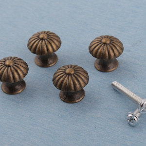 Może przedstawiać: Cztery antyczne mosiężne gałki do szafek z motywem słonecznika. Gałki są okrągłe z żłobionym wzorem i osadzone na krótkiej, zaokrąglonej podstawie. W zestawie znajdują się również dwie srebrne śruby.