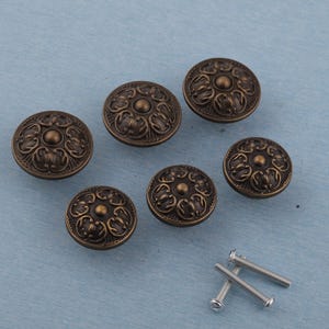 以下が含まれることがあります： 装飾的なフローラルディテールが施された、アンティークブロンズのキャビネットノブ6個セット。丸いノブは、渦巻き模様に囲まれた隆起した中央のデザインが特徴です。2本のシルバーネジも含まれています。