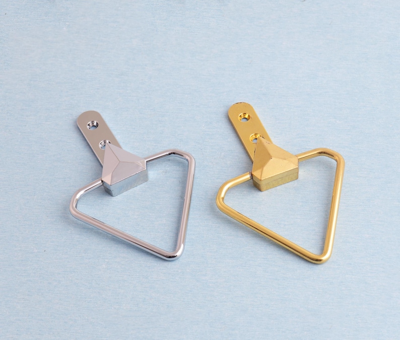 May include: Two metal cabinet pulls. One is silver, the other is gold. Each pull features a triangular handle and a rectangular base with a triangular accent. The pulls have a mounting plate with two holes.