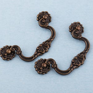May include: Two antique bronze drawer pulls with floral detailing. Each pull features a curved handle and decorative end pieces. The pulls are made of metal and have a vintage aesthetic, suitable for furniture restoration or adding a classic touch to cabinets.