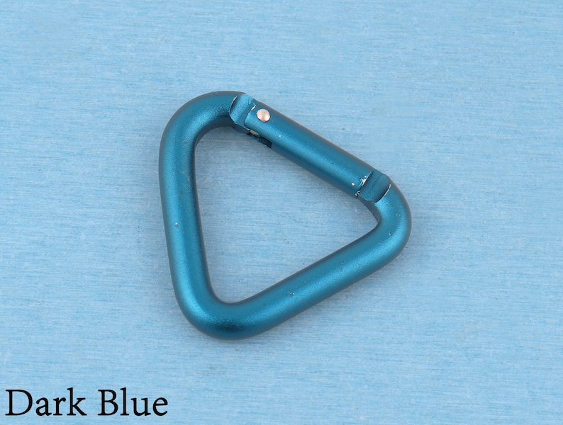 May include: A triangular carabiner with a metallic dark blue finish. The carabiner features a spring-loaded gate mechanism on one side, allowing it to open and close securely. The surface has a smooth, matte texture throughout the entire frame.