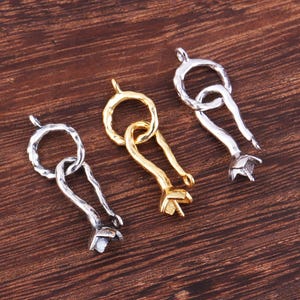 May include: Three metal jewellery clasps in silver and gold tones. Each clasp features a ring connected to a curved, textured body ending in a flower-like shape. The clasps are displayed on a dark wood surface.