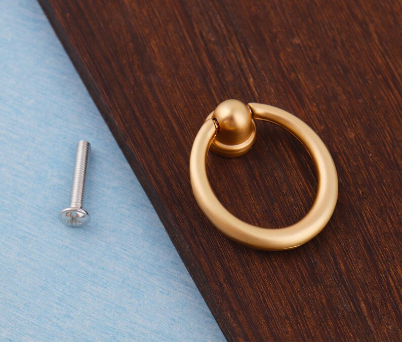 Op de afbeelding: Een goudkleurige ringtrekker met een ronde handgreep en een kleine, ronde basis. Een zilveren schroef bevindt zich links van de trekker. De hardware is geplaatst op een donkerbruin houten oppervlak en een lichtblauw oppervlak.