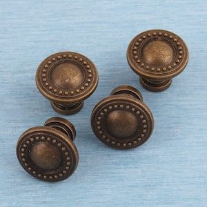 May include: Four antique bronze cabinet knobs with a circular design and a beaded edge. The knobs are round with a slightly domed center and a short stem. They are arranged on a light blue surface.
