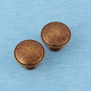 Peut inclure: Deux boutons de placard ronds avec une finition en laiton antique vieilli. Chaque bouton présente une surface supérieure lisse et légèrement bombée ainsi qu'une base circulaire, affichant un aspect métallique texturé et patiné.