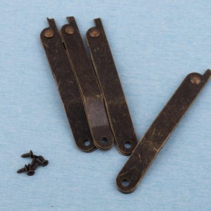 Op de afbeelding: Een verzameling antiek bronzen scharnieren en kleine schroeven. De scharnieren zijn rechthoekig met afgeronde uiteinden en een kleine, gebogen verlenging. Elk scharnier heeft een klinknagel. De schroeven zijn klein en donker.