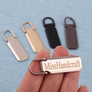 K&ouml;nnte beinhalten: Eine Hand h&auml;lt einen silbernen Metall-Rei&szlig;verschluss-Anh&auml;nger mit dem Text "MissHandcraft". Weitere Rei&szlig;verschluss-Anh&auml;nger in Gold, Schwarz und Braun sind im Hintergrund. Die Anh&auml;nger haben eine rechteckige Form mit einer Schlaufe.