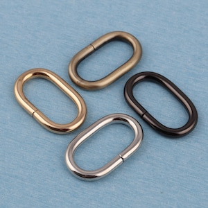 Op de afbeelding: Vier ovale metalen ringen in verschillende afwerkingen: goud, zilver, zwart en antiek messing. Deze ringen worden vaak gebruikt om banden of kettingen aan tassen, handtassen of andere accessoires te bevestigen.